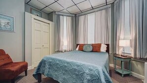 Un dormitori amb un llit, una cadira, una tauleta de nit i una finestra amb cortines.