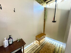Une salle de bain avec des murs carrelés de carreaux blancs, un banc en bois, une table en bois avec des articles de toilette et un espace douche avec un sol en bois.