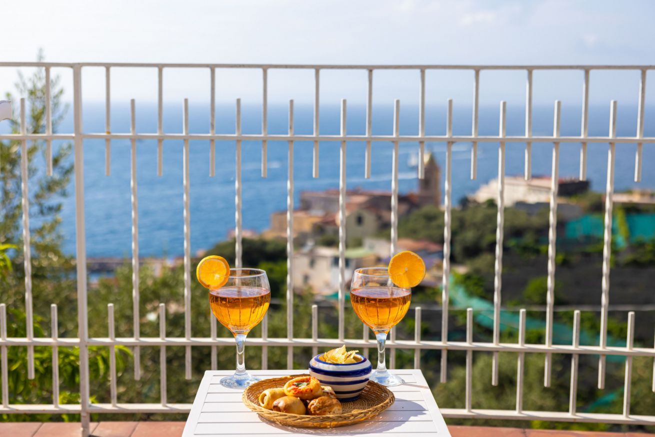 Un balcon avec une table dressée pour deux personnes, agrémentée de verres contenant un liquide ambré, d’un panier de viennoiseries et d’un panier en osier.