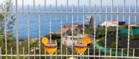 A balcony with a table set for two, featuring glasses of amber liquid, a bowl of pastries, and a wicker basket.