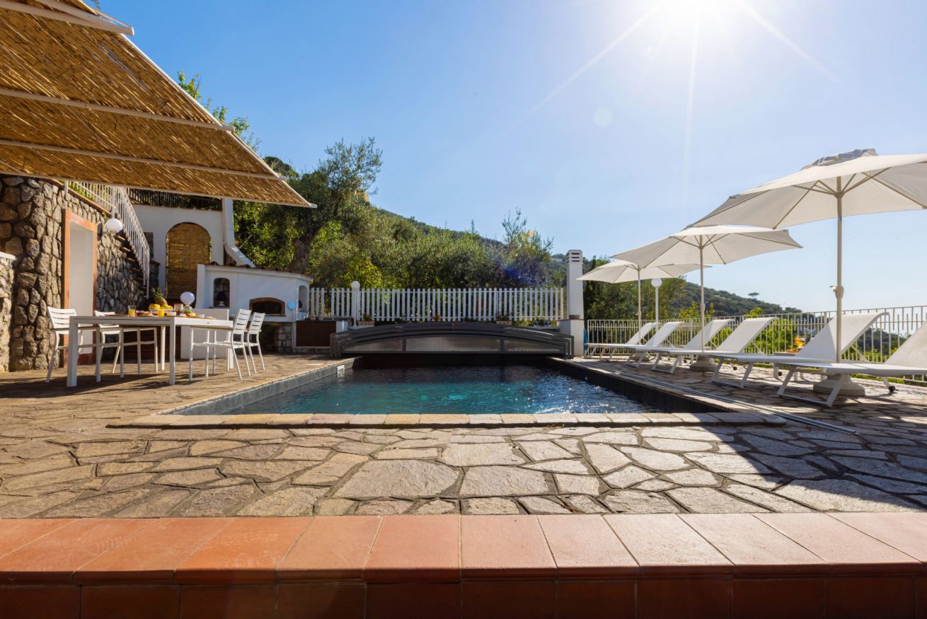 A pool area with stone flooring, a bridge, and lounge chairs with umbrellas.