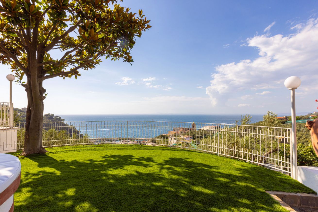 Un espacio al aire libre con césped, un árbol, una valla blanca y vista al mar.