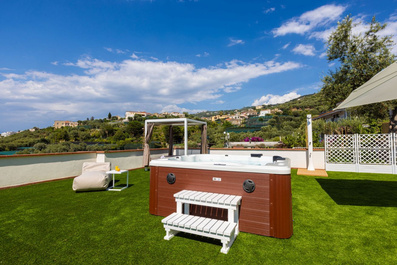 Un bain à remous avec une structure extérieure en bois et un banc blanc, installé dans un espace pelouse bien entretenu, avec une vue panoramique sur une colline en arrière-plan.