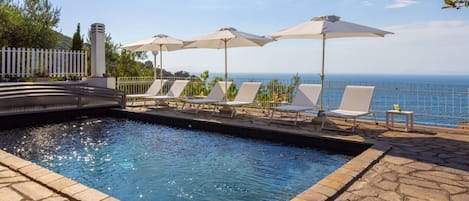 Zona de piscina con sillones de descanso y sombrillas, un borde de piedra y vistas al mar.