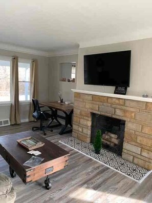 A living room with a fireplace, a television mounted above it, a dining area with a table and chairs, and a window with curtains. - Brick Ranch On A Quiet Street In Historic Bedford (Bedford)