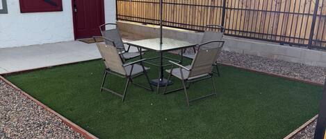 Un patio con mesa y sillas, rodeado de césped artificial y un borde de grava.