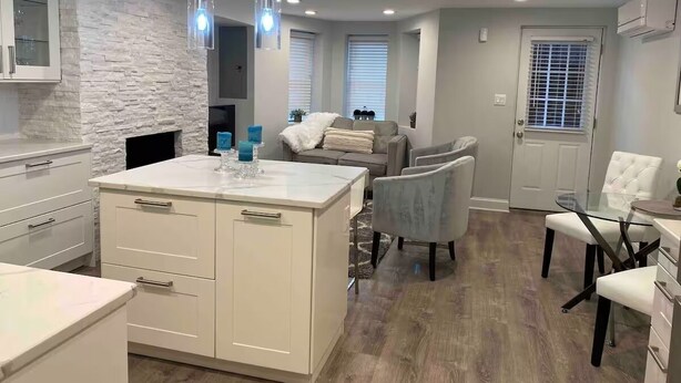 Una cocina moderna con armarios blancos, una isla central e iluminación colgante. Cuenta con un área de estar con sofá y sillones.