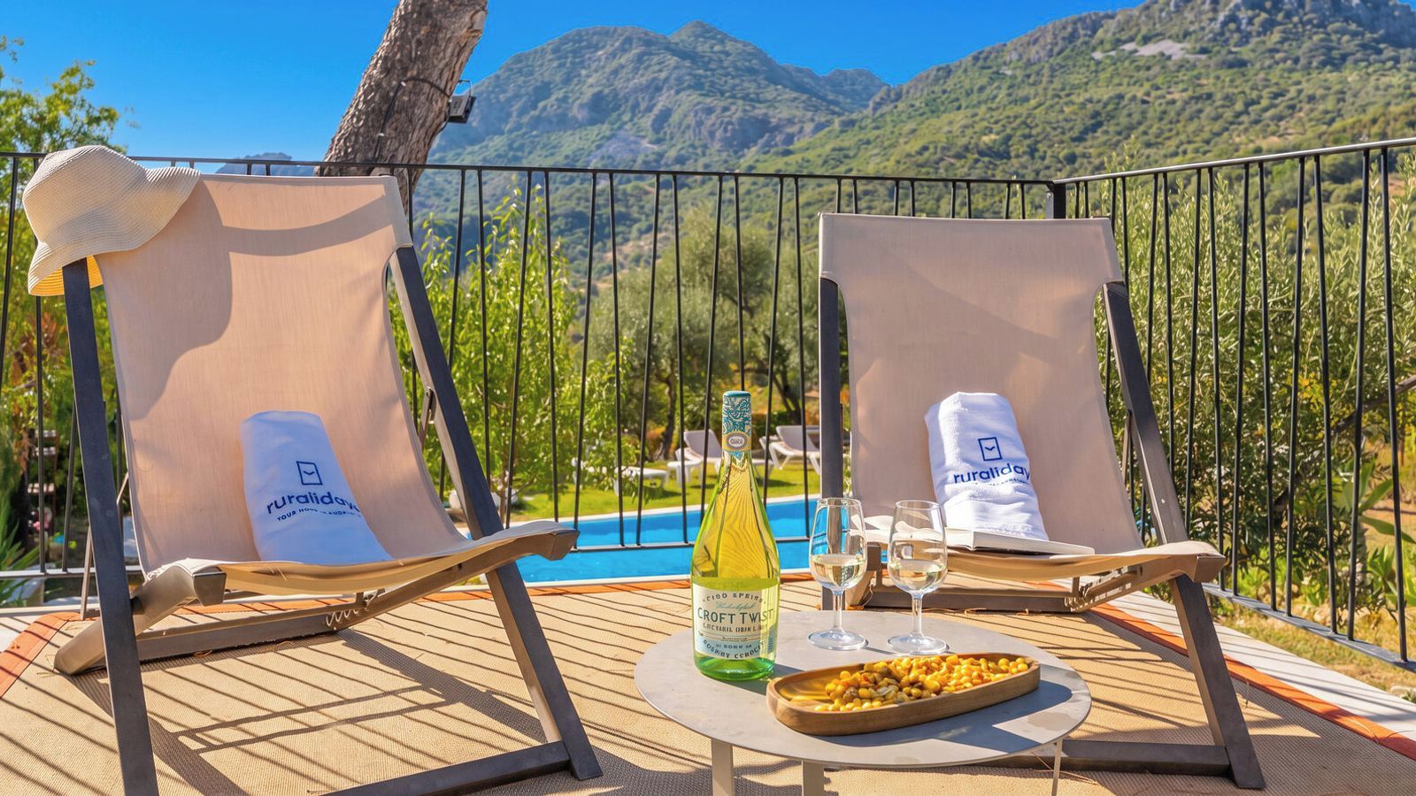 Deux fauteuils de relaxation avec des serviettes, une bouteille de vin, des verres et un plateau de nourriture sur une table, le tout donnant sur une piscine et des montagnes.