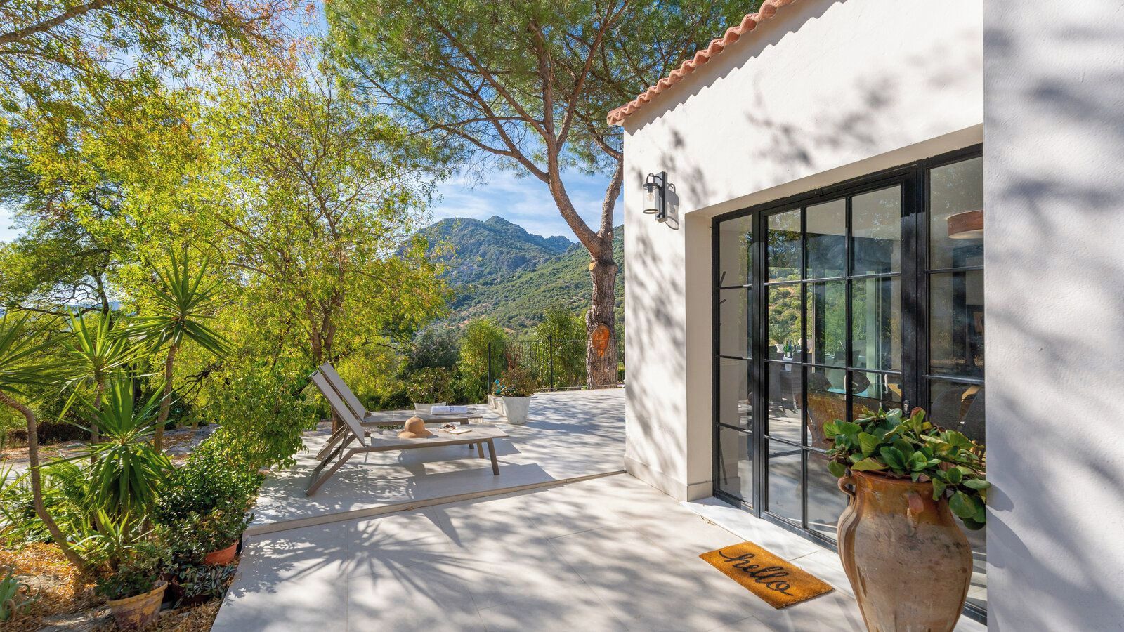 Une terrasse ensoleillée avec un bâtiment blanc, une porte vitrée et une vue sur les montagnes.