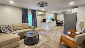 Un salón moderno con un sofá, una mesa de centro y sillones de mimbre. Se ve un área de cocina con refrigerador, microondas y una mesa de comedor al fondo.