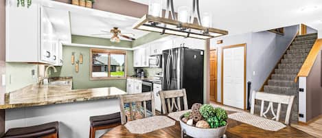 A modern kitchen with a central island, wooden chairs, a ceiling fan, and a staircase.