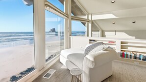 Una habitación moderna con un amplio ventanal con vistas a la playa, un sofá blanco, un taburete pequeño y una estantería.