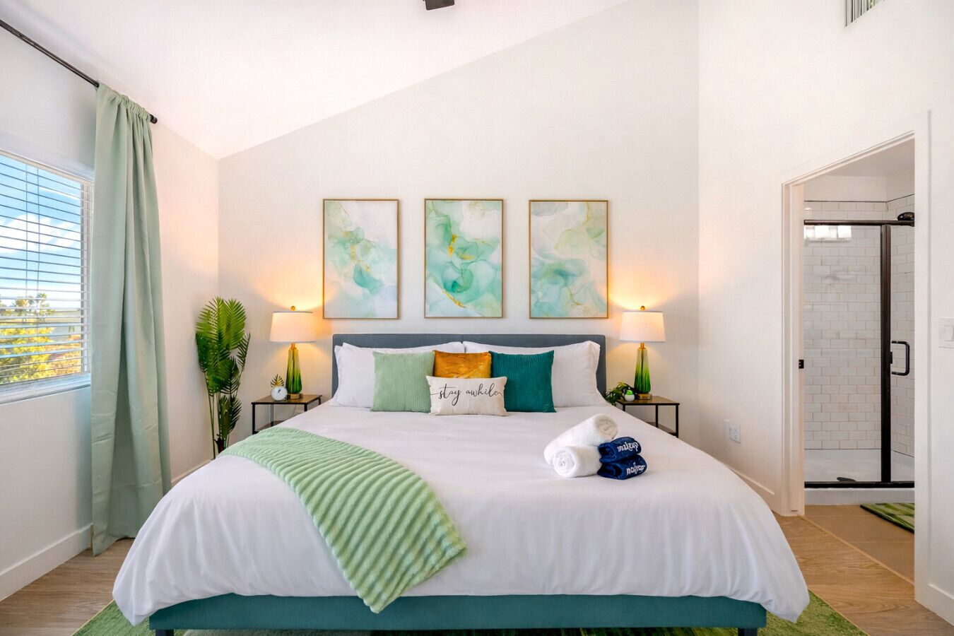 A hotel room with a bed, two bedside lamps, a window with curtains, and three abstract paintings on the wall.