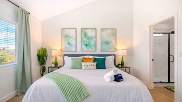 A hotel room with a bed, two bedside lamps, a window with curtains, and three abstract paintings on the wall.