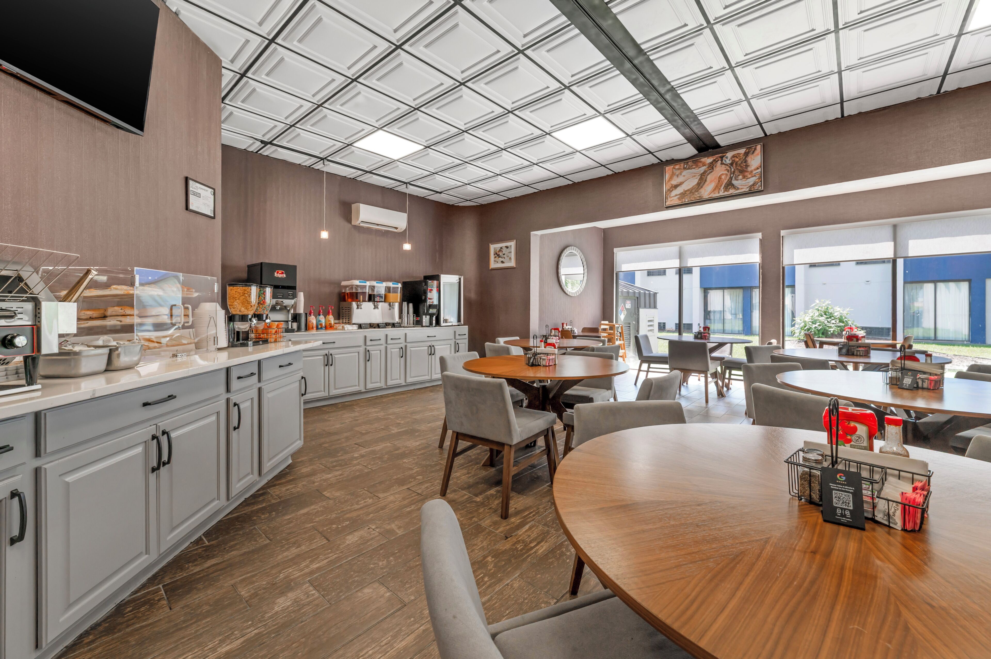 A modern kitchen with a large island, a dining area with round tables, and a view of the outdoors through large windows.