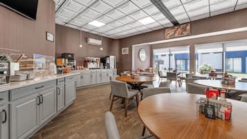 A modern kitchen with a large island, a dining area with round tables, and a view of the outdoors through large windows.
