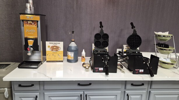 A kitchen counter with a waffle maker, a bottle of waffle batter, and a box of fresh waffles. - Red Lion Inn & Suites Kansas City (Kansas City)