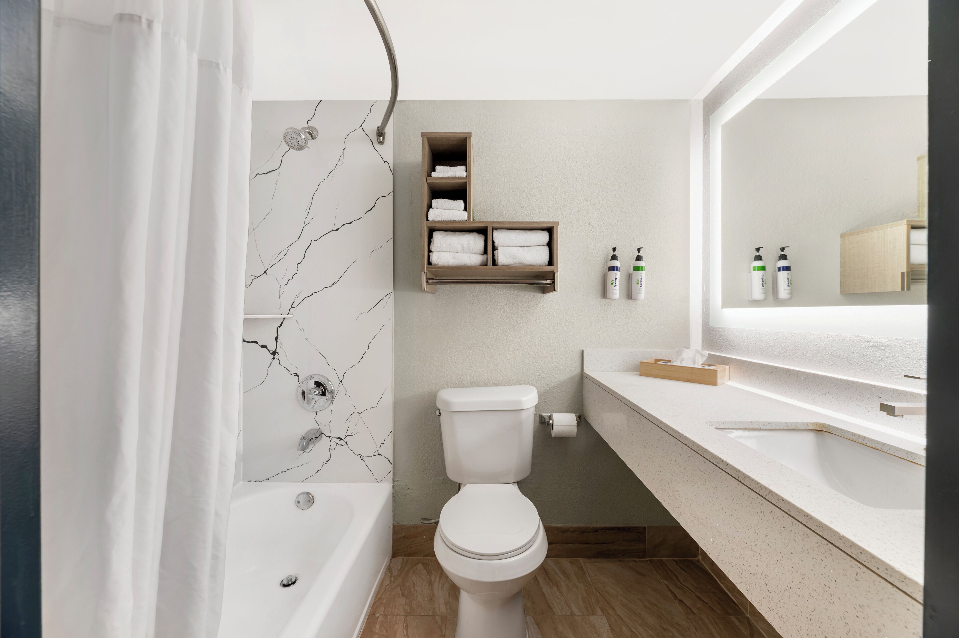 A modern bathroom with a marble wall, a bathtub, a toilet, a vanity with a mirror, and shelves with toiletries.