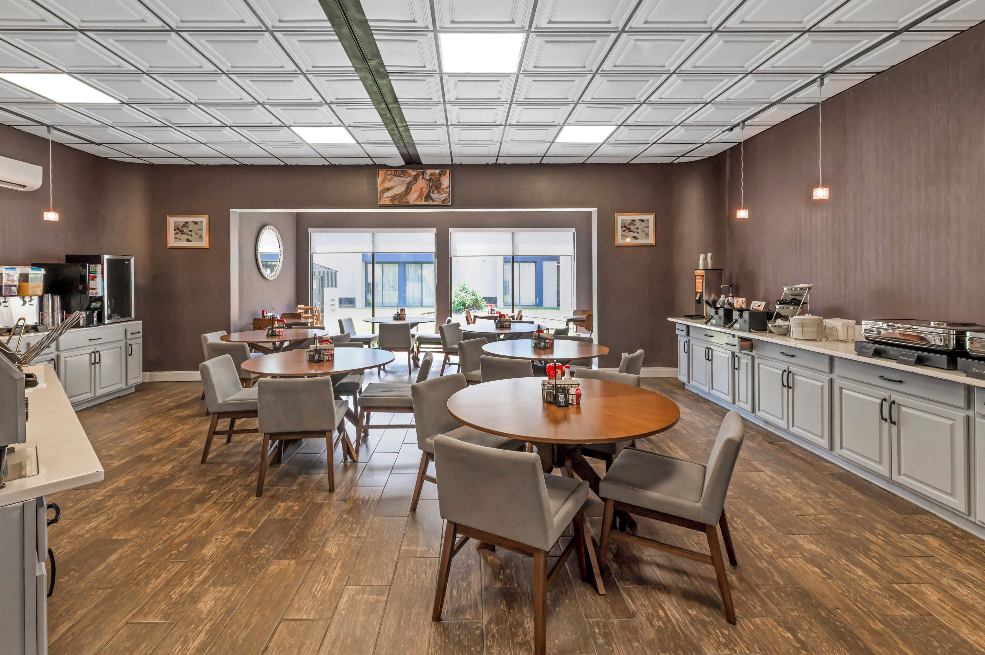 A spacious dining area with multiple round tables and chairs, a kitchen area with modern appliances, and large windows providing natural light.