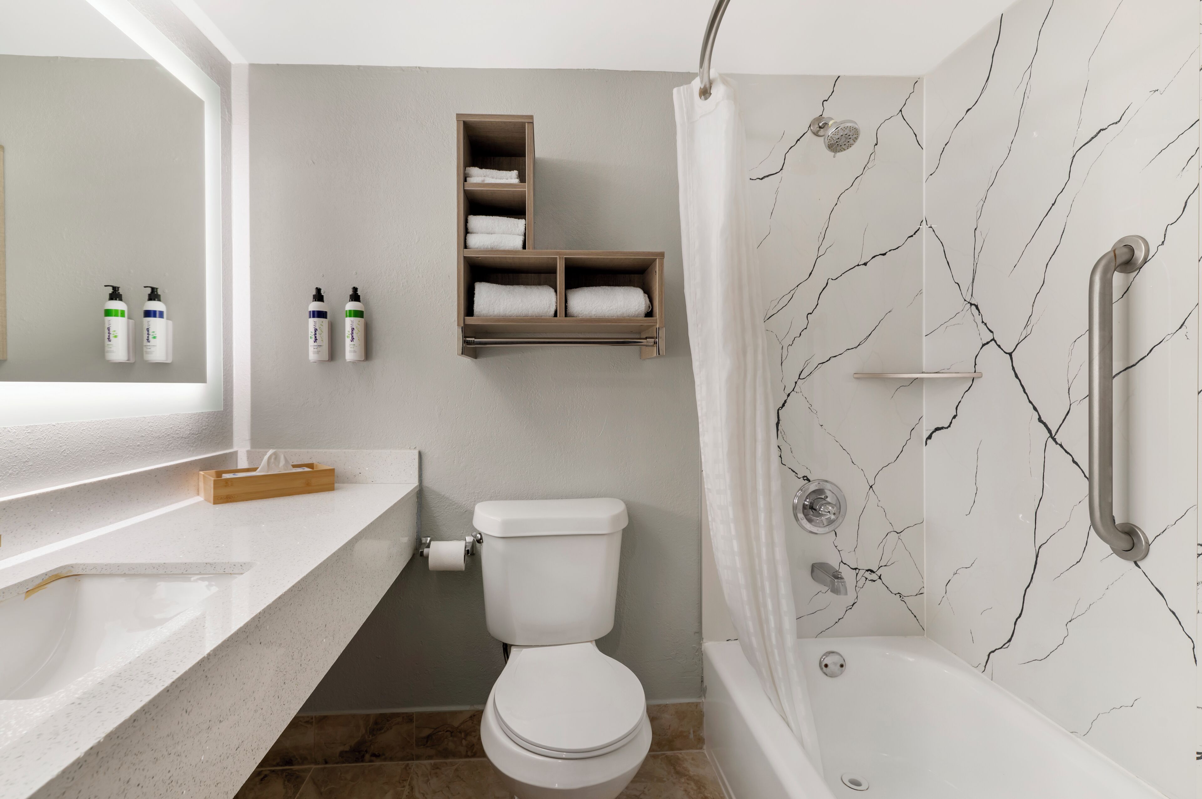 A modern bathroom with a white toilet, a shower with a grab bar, a marble-patterned wall, and a wooden shelf with towels.