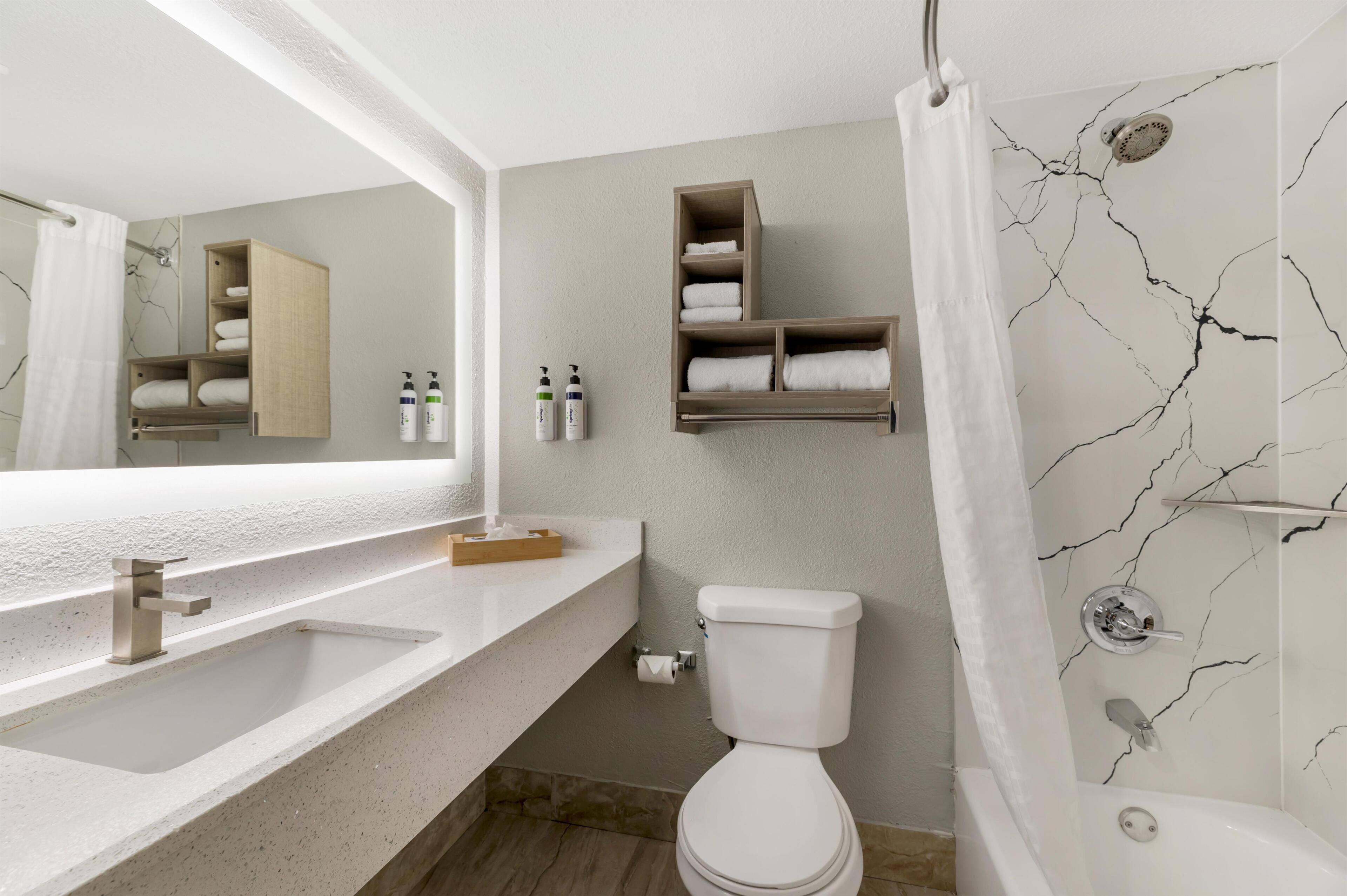 A bathroom with a white sink, toilet, and bathtub, marble-patterned walls, and wooden shelves with towels.