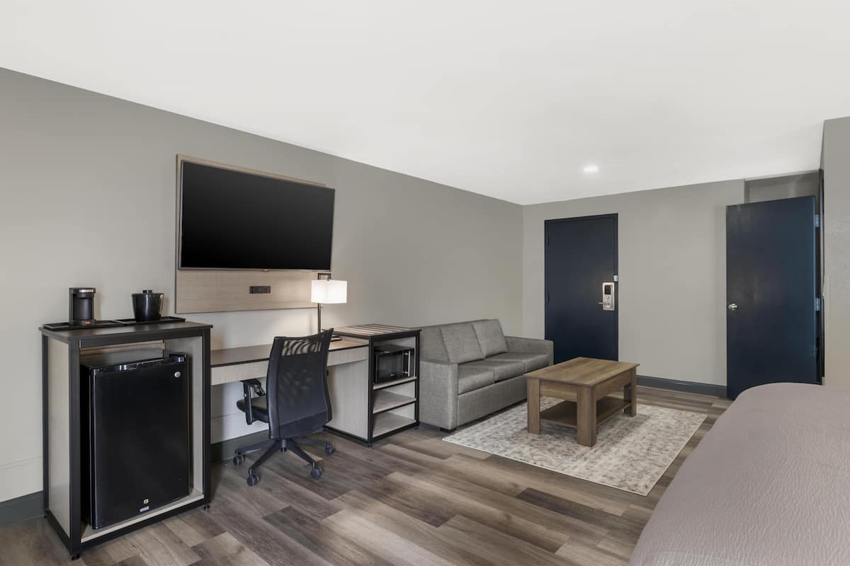 A hotel room with a desk, a sofa, a TV, a mini fridge, a lamp, and a bed.