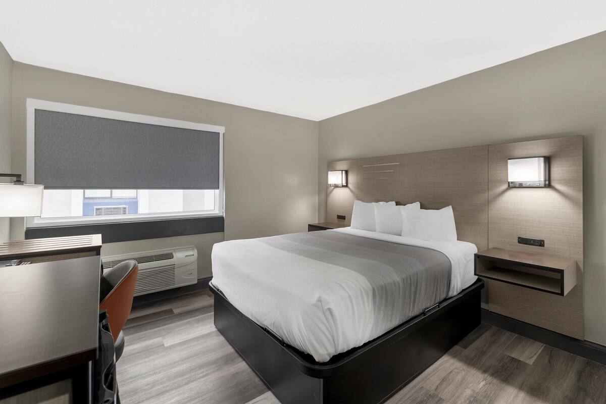 A modern hotel room with a bed, desk, chair, and a large window.
