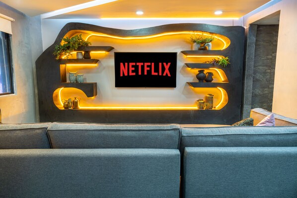 A modern living room with a large flat-screen TV mounted on a wall, surrounded by shelves with decorative items and plants. - 5 bedroom Apartment with Massive Jacuzzi and Office space (Medellín)