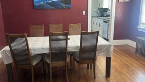 A dining area with a wooden table and chairs, a painting on the wall, and a kitchen in the background.