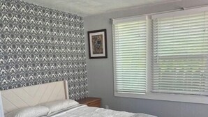 A bedroom with a bed, a patterned wallpaper, a window with blinds, and a framed picture on the wall.