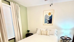 Een net opgemaakt bed in een kamer met een plafondventilator, een klein tafeltje met een lamp en een schilderij aan de muur.