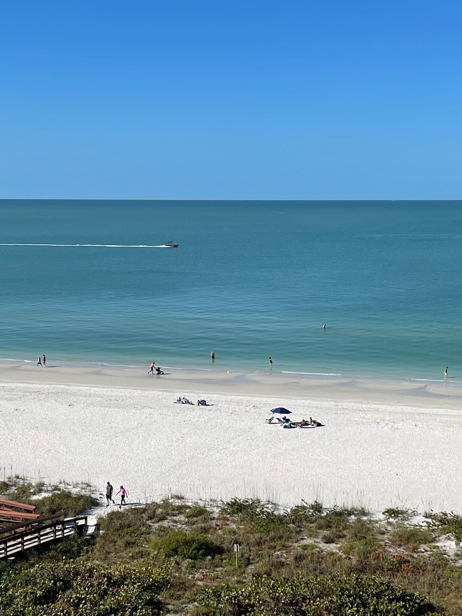 Ein Strand mit blauem Himmel, ruhigem Wasser und einigen Personen, die am Ufer entlanggehen.
