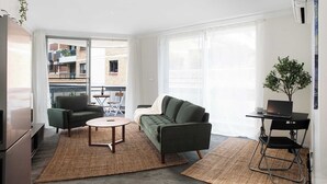 Una sala de estar moderna con sofá, sillón y mesa de centro, una laptop sobre un escritorio, y vista al balcón exterior.