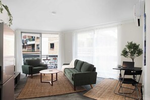 Ein modernes Wohnzimmer mit einer Couch, einem Sessel und einem Couchtisch, einem Laptop auf einem Schreibtisch sowie Blick auf den Balkon.