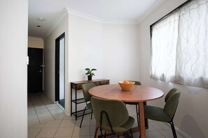 A dining area with a round table, four chairs, a bowl of fruit, a potted plant, and a window with curtains. - Urban Oasis 2BR with Huge Deck & Secure Parking (Potts Point)