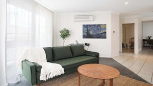 Una sala de estar moderna con un sofá verde, una mesa redonda de madera, un cuadro en la pared y una planta.