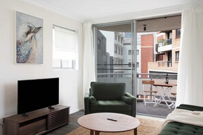 En moderne stue med et fladskærms-tv, en grøn lænestol, et rundt sofabord og en skydedør, der fører ud til en balkon med havemøbler.