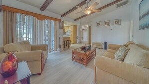 Uma sala de estar espaçosa com teto alto, vigas de madeira, ventilador de teto e cozinha ao fundo.