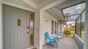 Varanda coberta com uma porta cinzenta, duas cadeiras azuis e vista para um jardim.