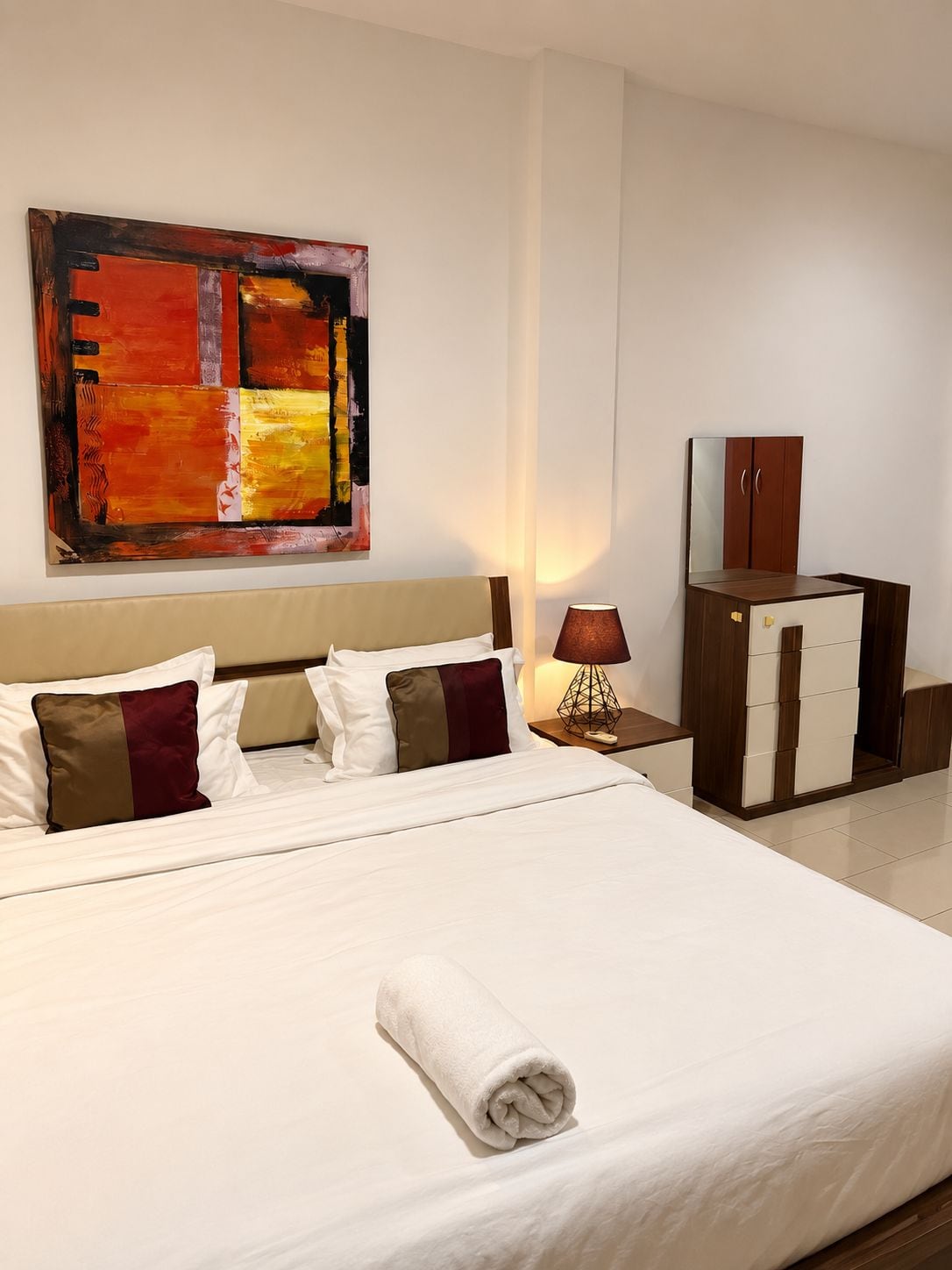 A hotel room with a large bed, a nightstand, a lamp, a wardrobe, and a painting on the wall.