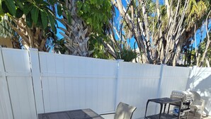 Un patio exterior con una mesa y sillas de metal, una parrilla y una cerca blanca.