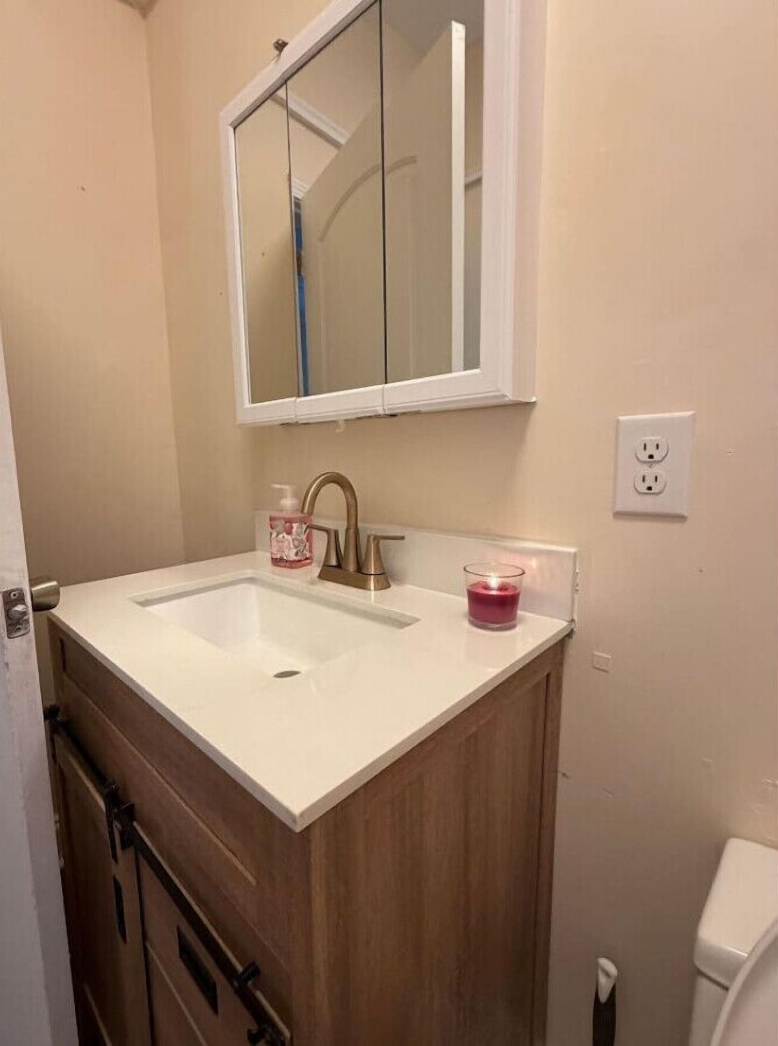 Een badkamer met een wit wastafel, een spiegel erboven, een houten kastje eronder, en een toilet aan de rechterkant.