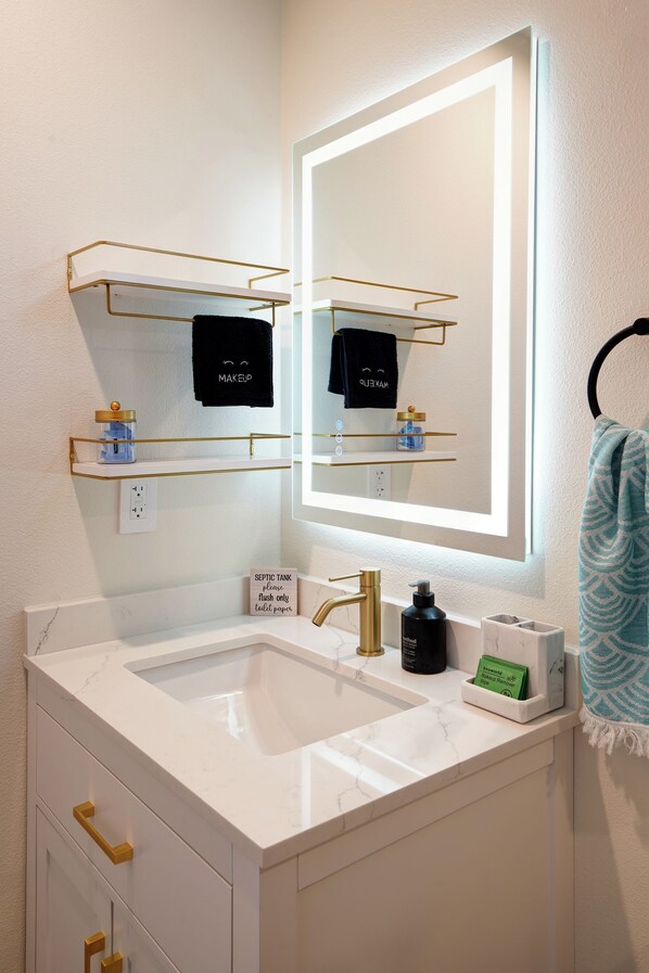 Un baño moderno con un lavamanos blanco, accesorios dorados y un espejo con iluminación integrada. Hay dos repisas sobre el lavamanos, una con un neceser y la otra con una caja de pañuelos. Un letrero en la pared indica que el inodoro es solo para desechos.