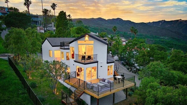 A modern two-story house with a balcony, surrounded by lush greenery and mountains in the background.