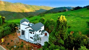A white house with a large roof, surrounded by lush green hills and a clear sky.