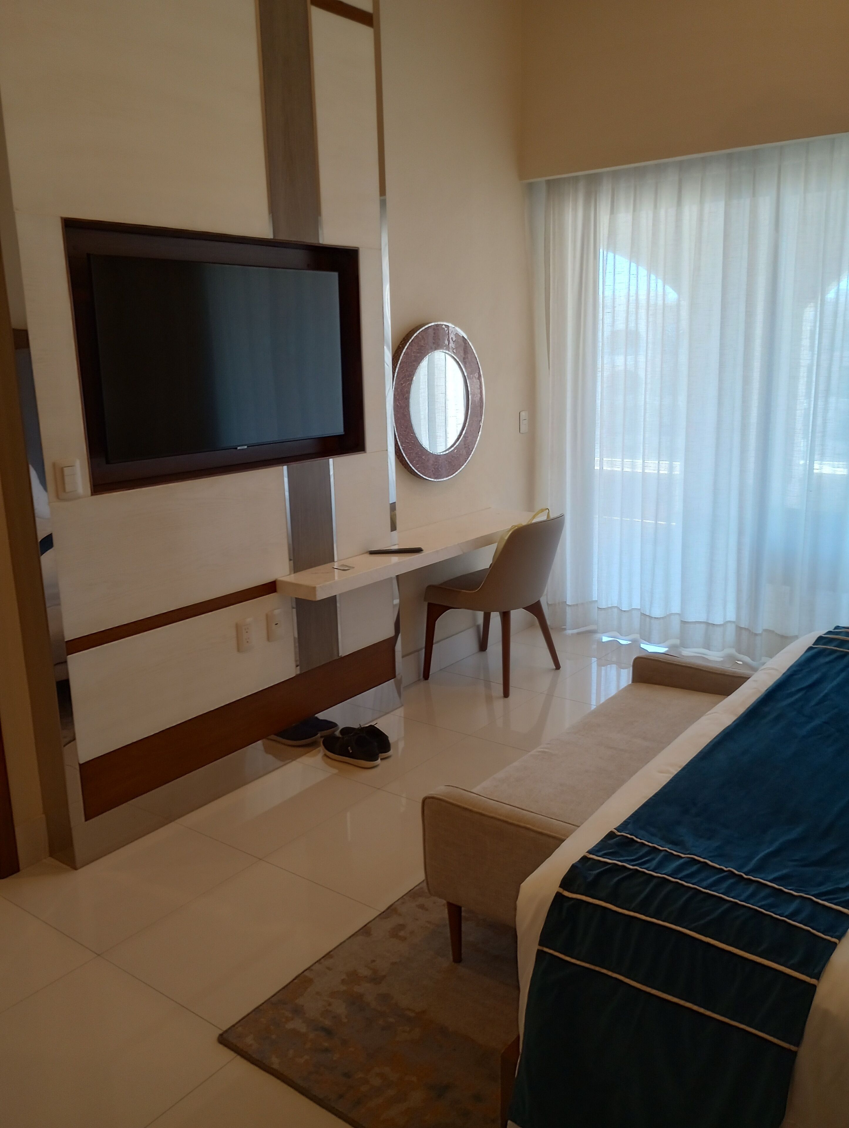 A hotel room with a bed, a desk, a television, a mirror, and a view of the outdoors through sheer curtains.