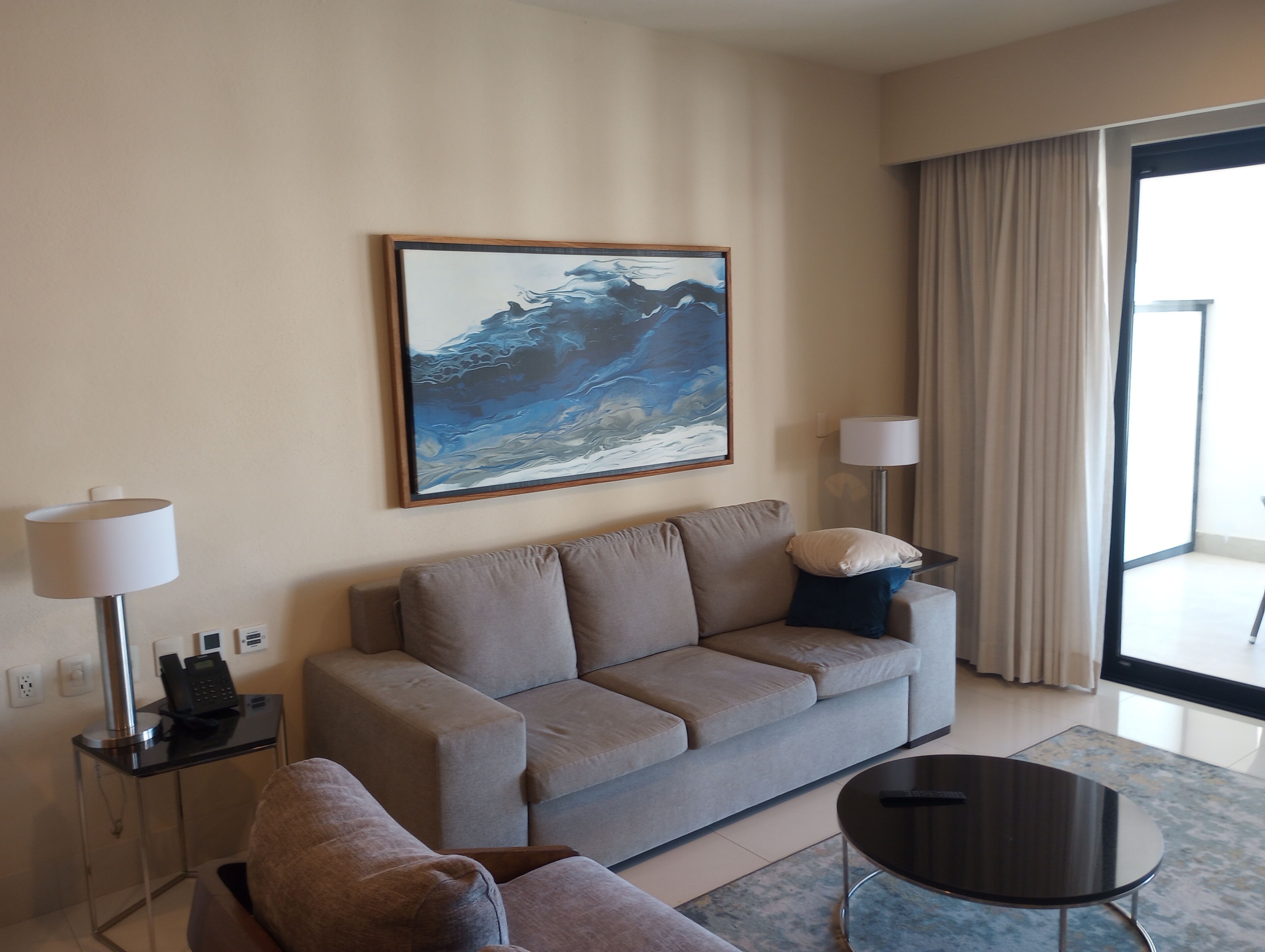 Et moderne hotelværelse med en beige sofa, et lille rundt bord, en lampe og et stort abstrakt billede på væggen.