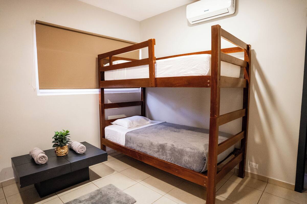 A bunk bed with a wooden frame, a small bedside table with a plant, and a window with a blind.