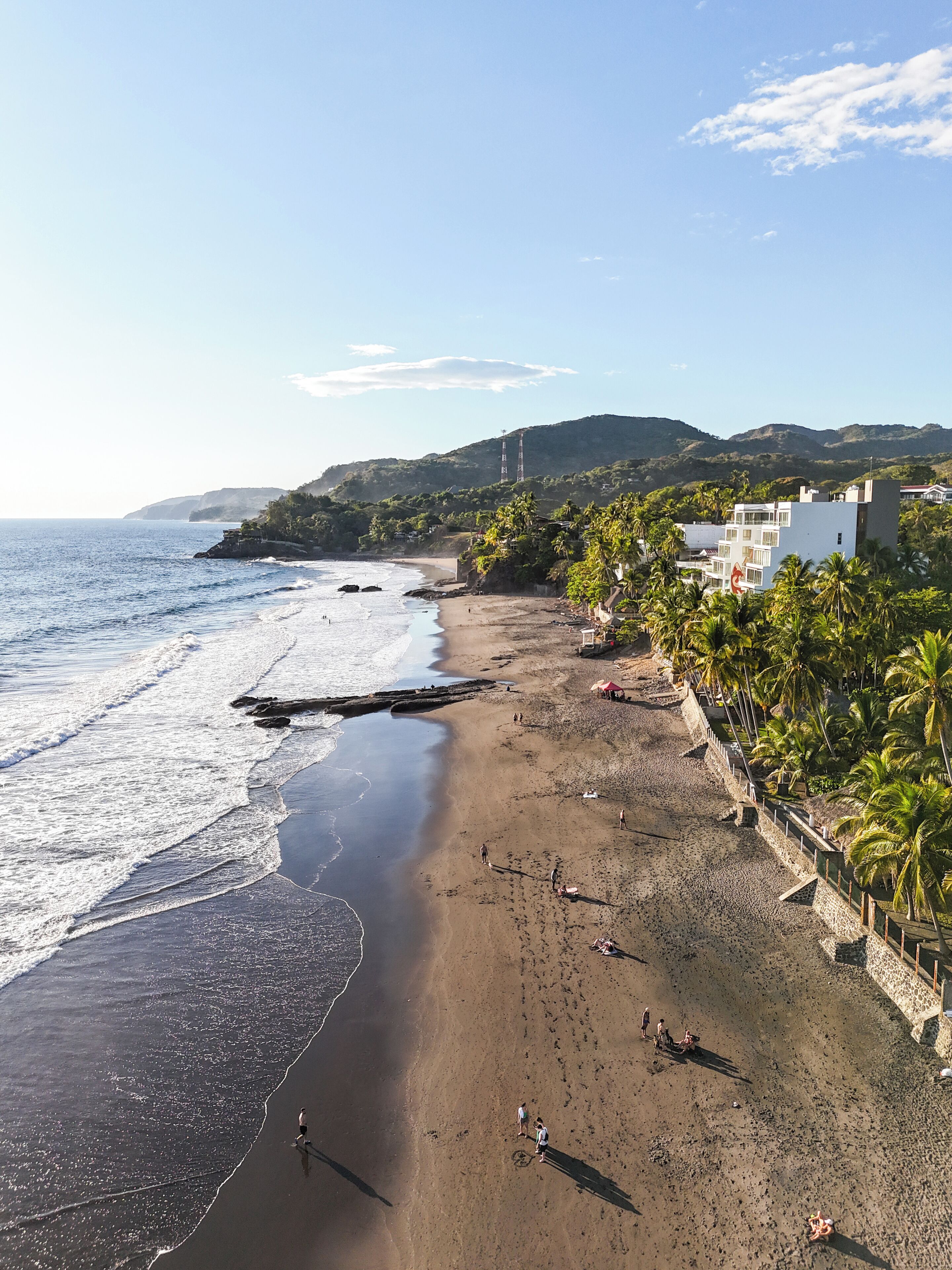 Una playa con costa rocosa, palmeras y edificios al fondo.