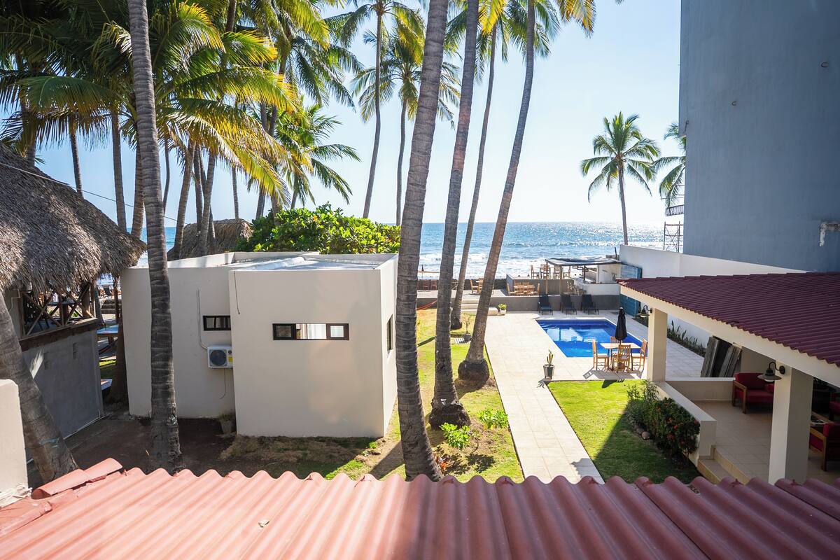 A pool area with a view of the ocean, surrounded by palm trees and a building with a red roof.